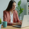 A young woman with long, straight brown hair sits at a light wooden desk, focused on her silver laptop. She is wearing a salmon-colored blazer over a white blouse. On her desk sits a teal coffee mug, a small potted plant, and colorful sticky notes. The background features a bright, organized office with another workstation, a large green potted plant, and a white shelving unit filled with office supplies.